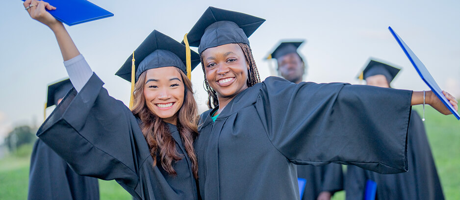Two smiling graduates in caps and gowns celebrating outdoors with arms raised, holding blue diploma folders.
