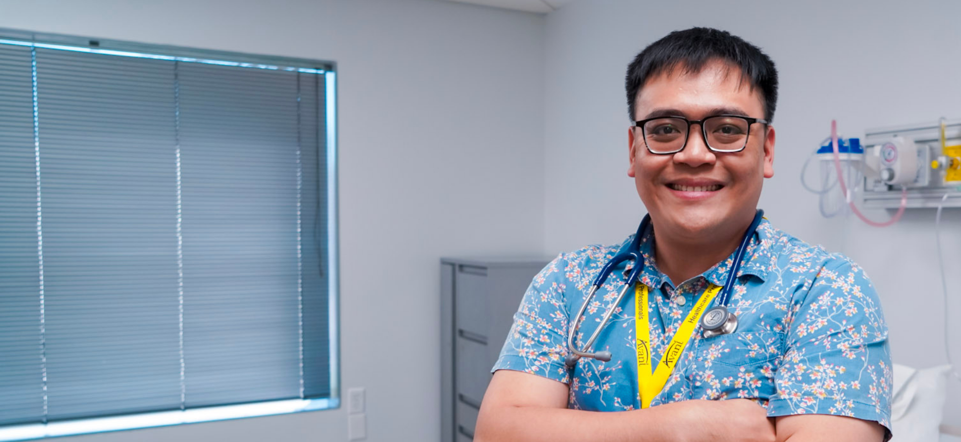 A smiling male healthcare professional wearing glasses, a stethoscope, and a floral shirt in a medical clinic.