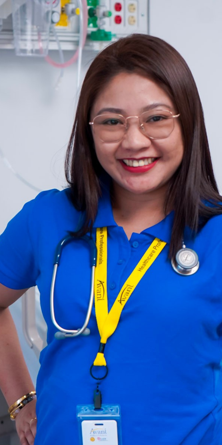 A smiling healthcare professional wearing glasses, a blue uniform, and a stethoscope, with an ID badge and yellow lanyard.