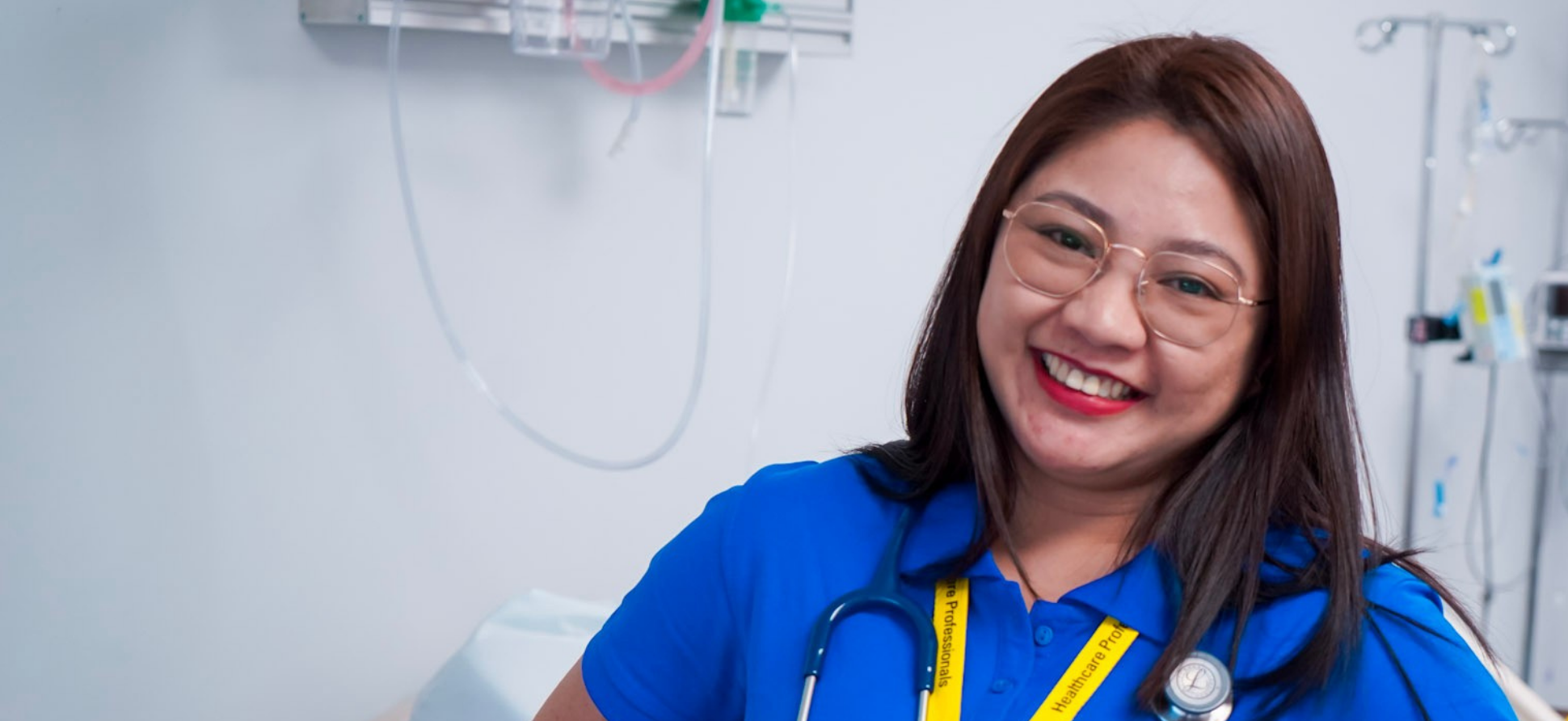 A smiling female healthcare professional with glasses and a stethoscope around her neck in a medical setting.
