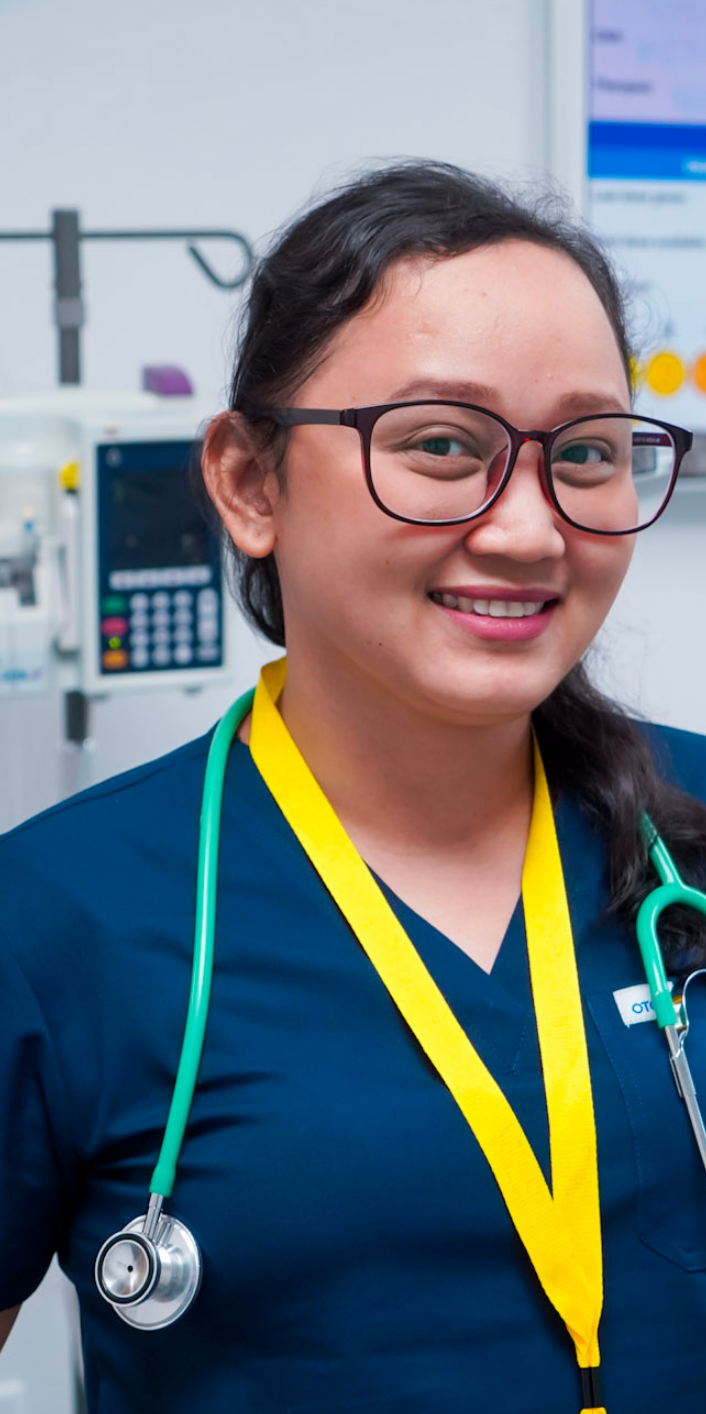 A smiling female healthcare professional wearing glasses, a stethoscope, and a yellow lanyard in a medical setting.