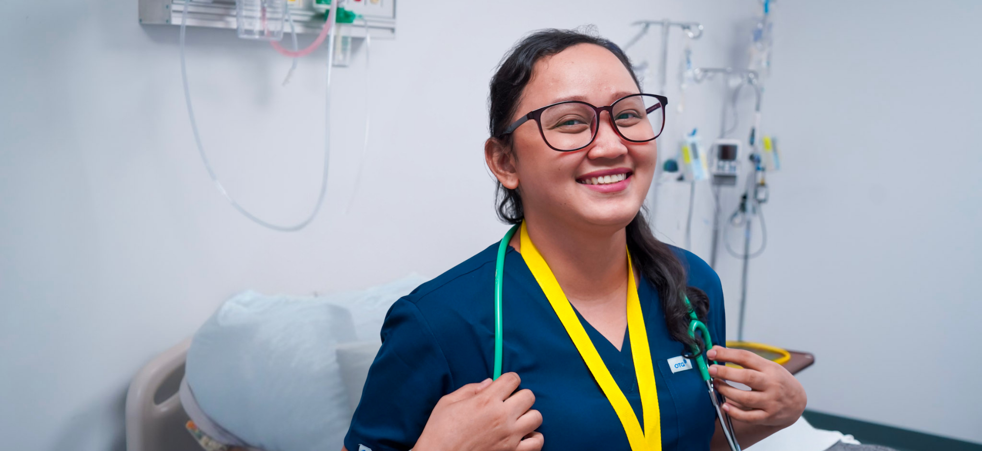 A smiling female healthcare professional wearing glasses, a stethoscope, and a navy blue uniform in a hospital room.