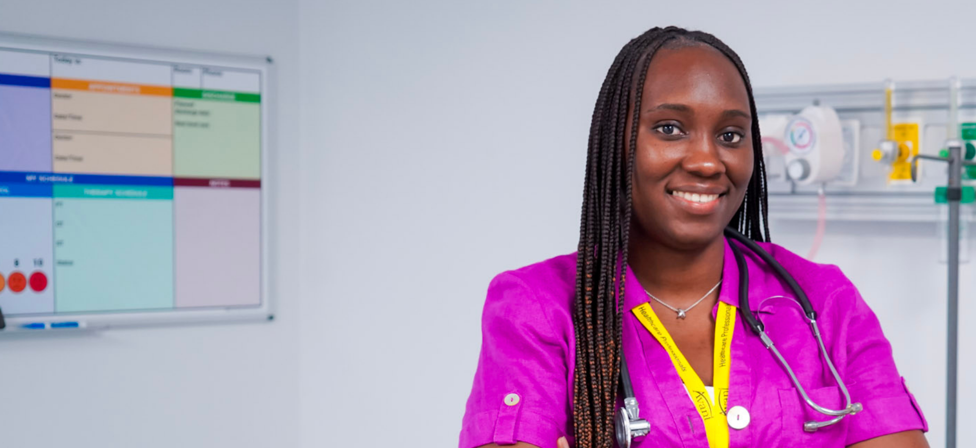 A smiling healthcare professional with braids, wearing a pink uniform and stethoscope, stands in a medical setting with