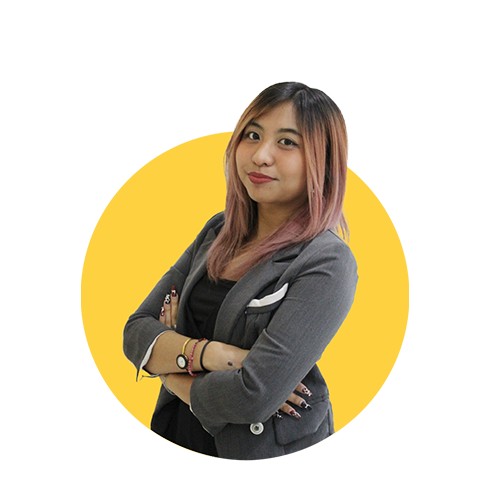 A confident woman with pink-tinted hair and a gray blazer crosses her arms against a yellow background.