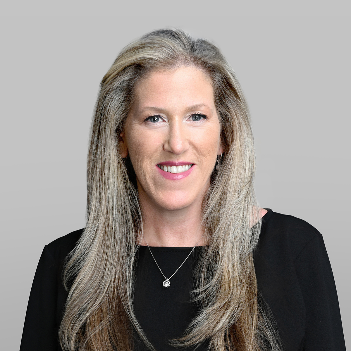 A woman with long, wavy gray-blonde hair, smiling, wearing a black top and a silver necklace against a gray background.