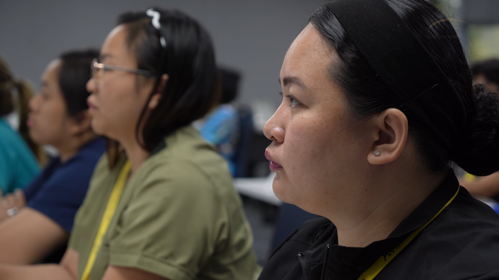 Close-up of a woman with dark hair in a bun, wearing earrings, attentively listening during a class or seminar.