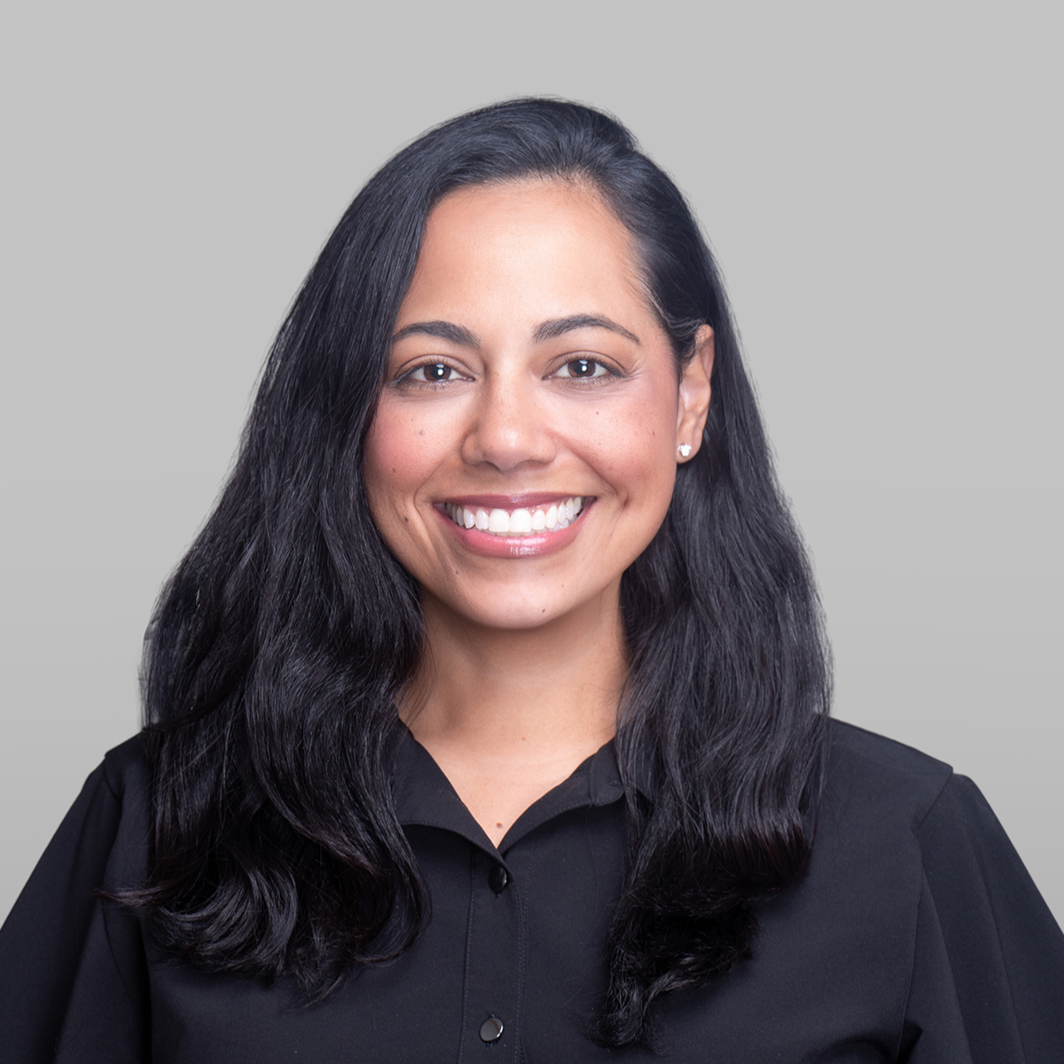 Cynthia Martinez-Patin has long, dark wavy hair, a bright smile, and wears a black collared shirt against a gray background.