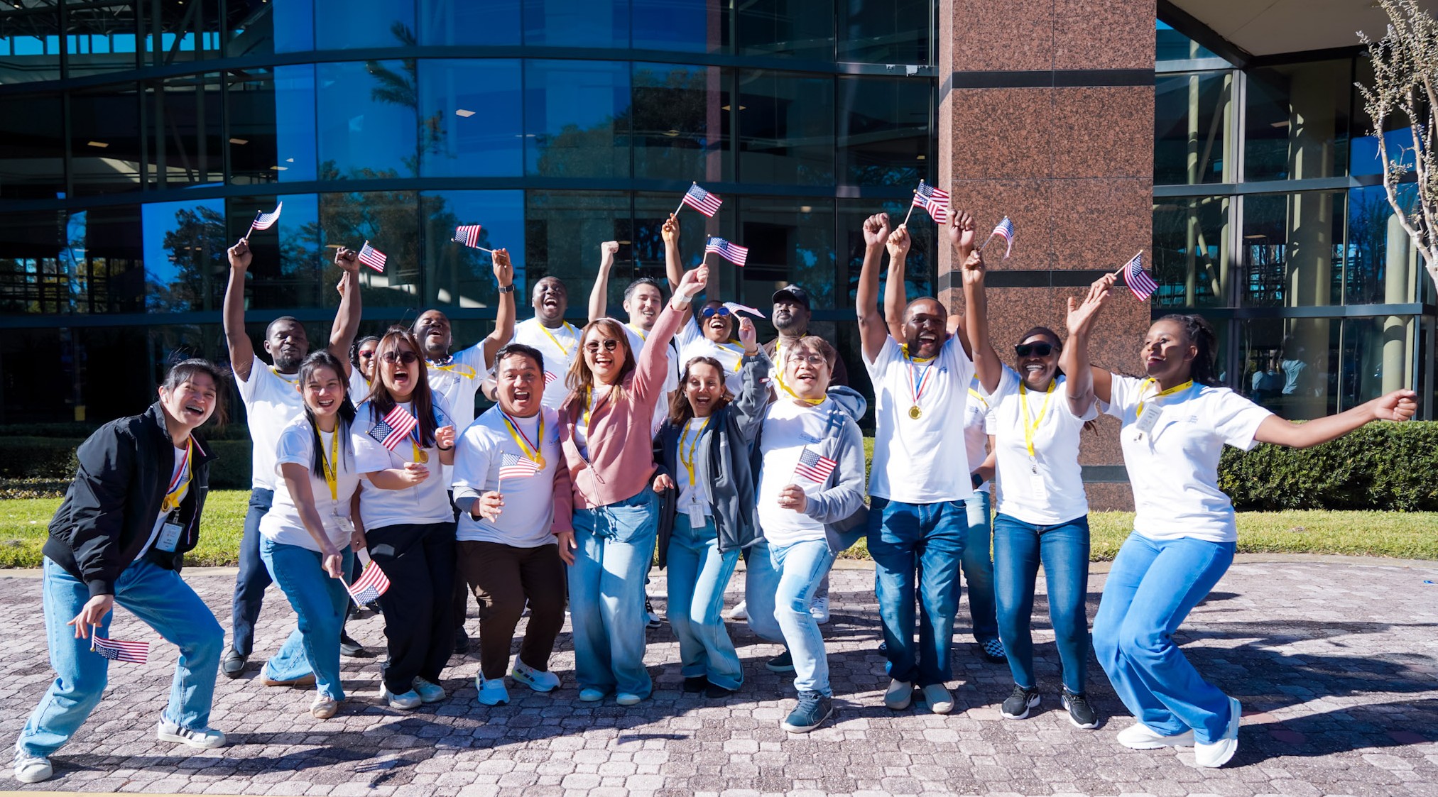 Group of diverse people celebrating outdoors with American flags in front of a modern glass building.