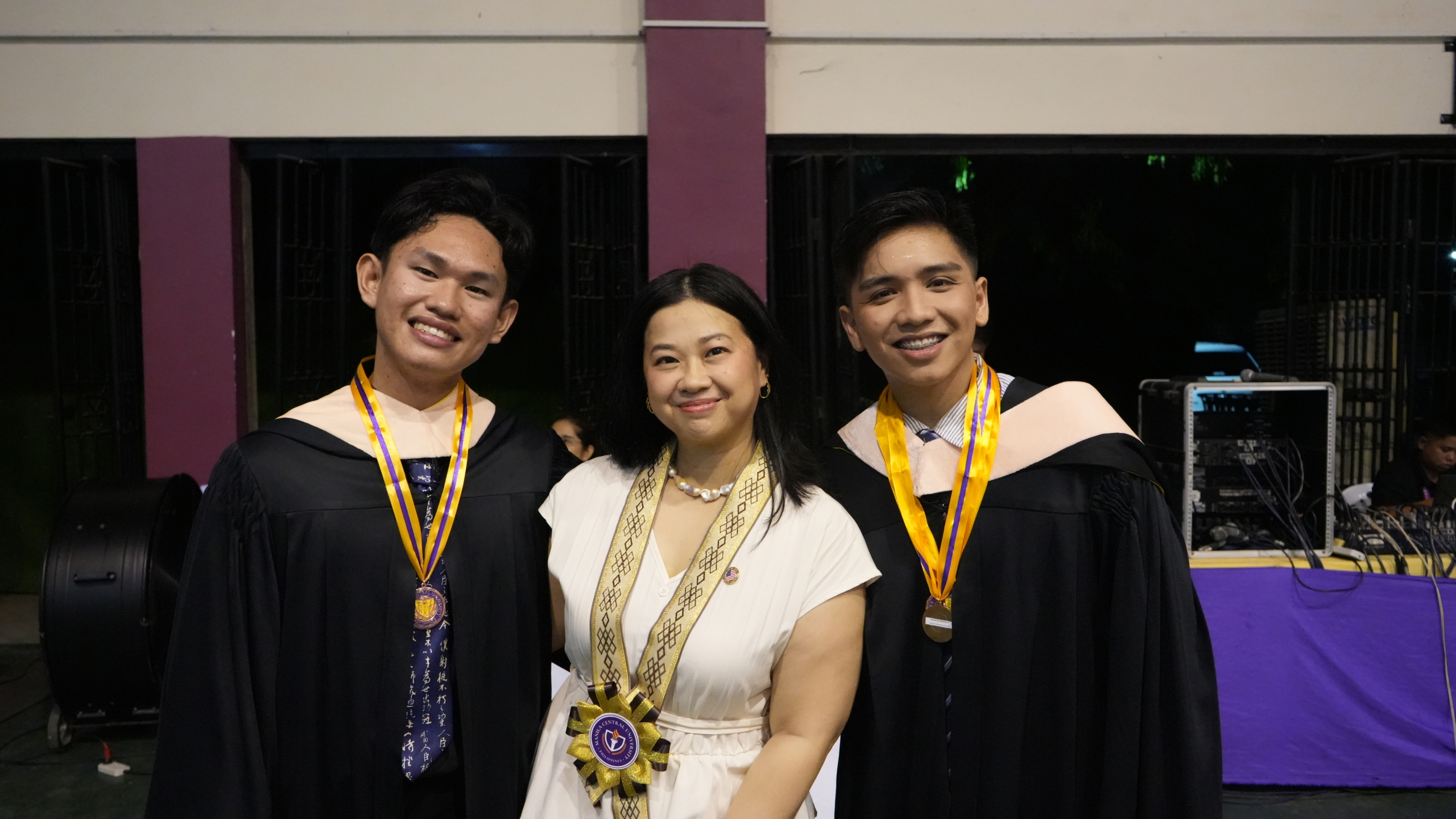 Two graduates in caps and gowns stand with a woman, all smiling, at a graduation ceremony.