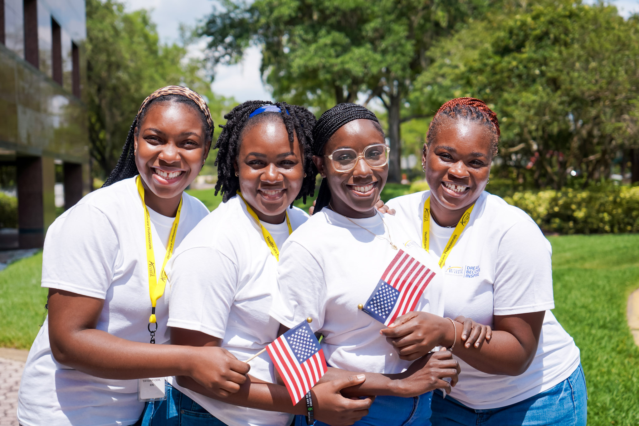 Four women smiling outdoors, holding small American flags, wearing white shirts and yellow lanyards.