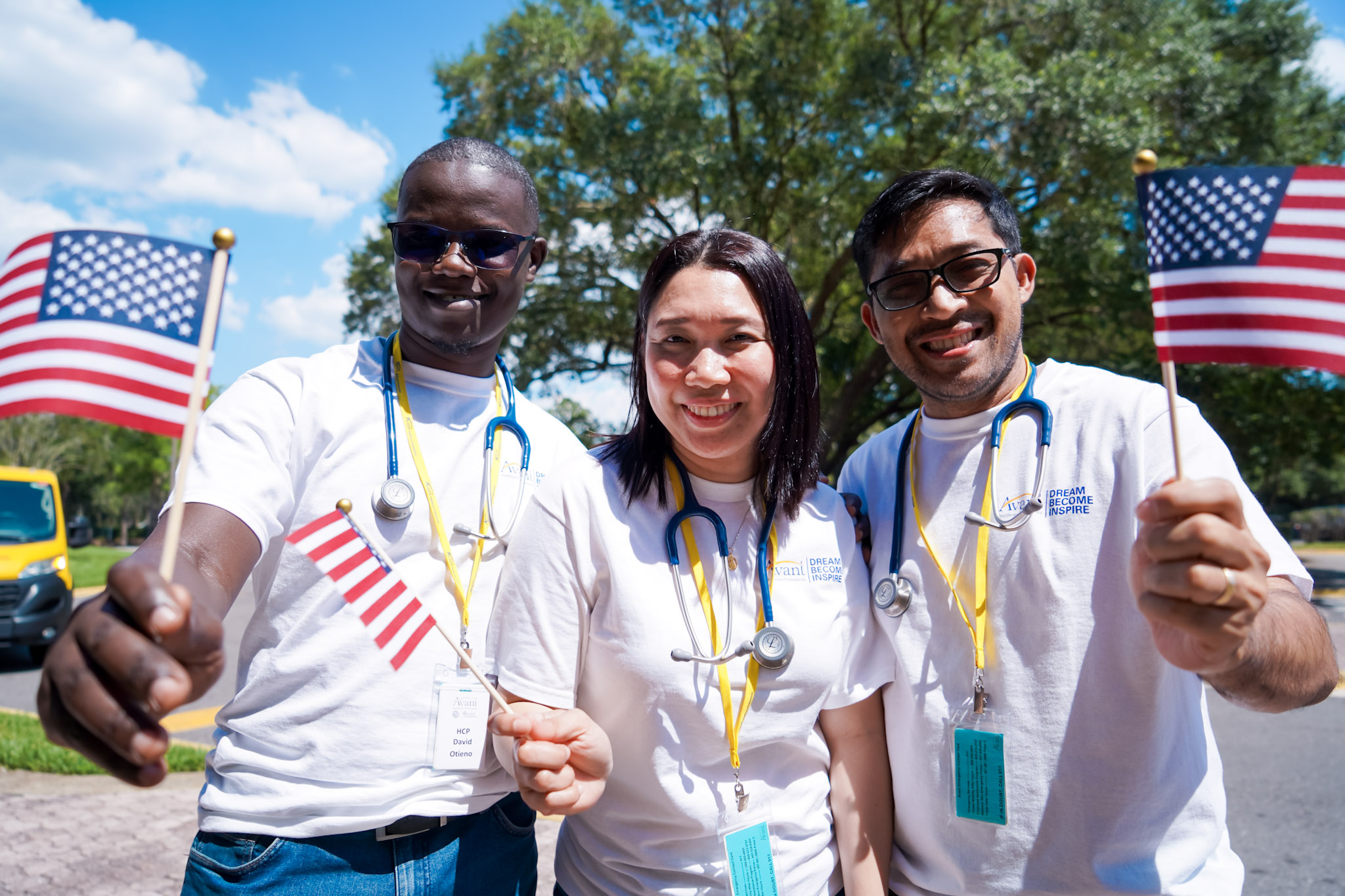 Three healthcare professionals smiling and holding small American flags outdoors on a sunny day.