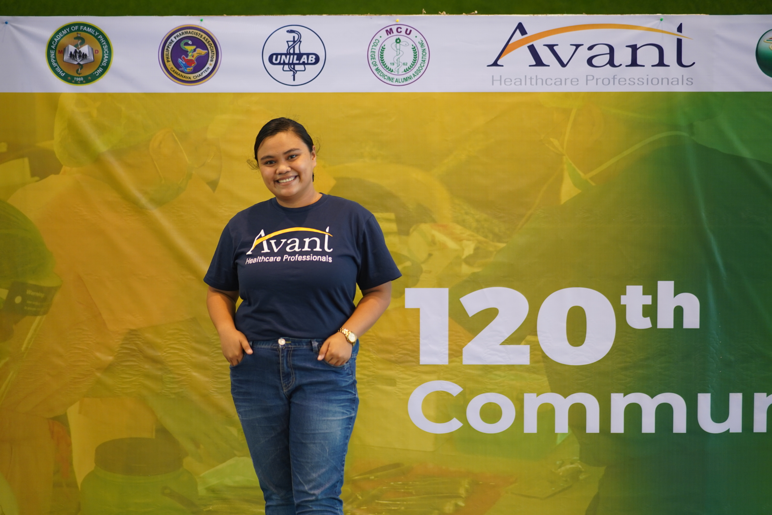 A smiling woman in a navy "Avani Healthcare Professionals" t-shirt stands in front of a yellow and green banner celebrating