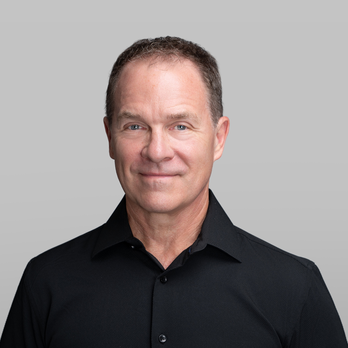 A man with short, dark hair and blue eyes wearing a black collared shirt against a gray background.