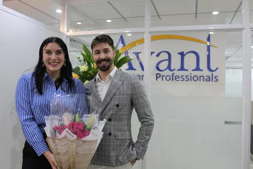A woman and a man stand together in an office, holding a bouquet of flowers, with a sign reading "Avanti Professionals"