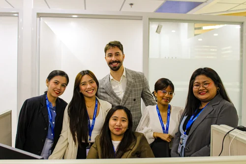 Five diverse professionals, four women and one man, smile together in an office setting.