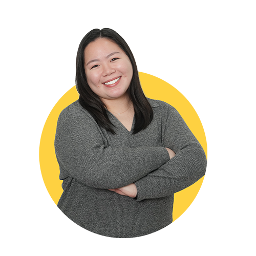 A smiling woman with long black hair, wearing a gray sweater, crosses her arms against a yellow background.