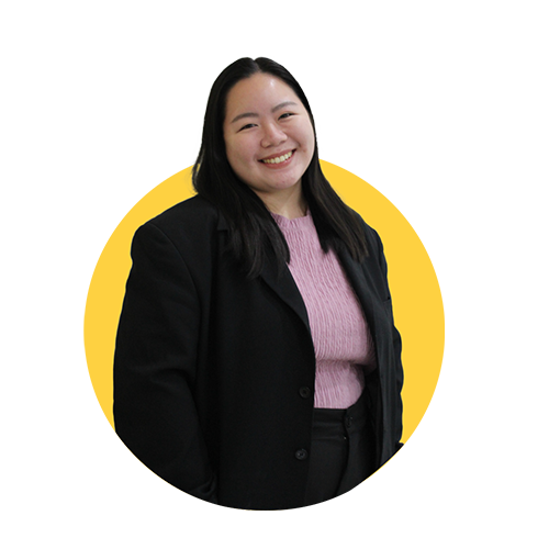 A smiling woman with long black hair wearing a black blazer over a pink sweater, standing against a yellow background.