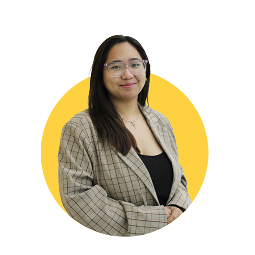 A woman with glasses and long dark hair smiling, wearing a plaid blazer over a black top, standing against a yellow