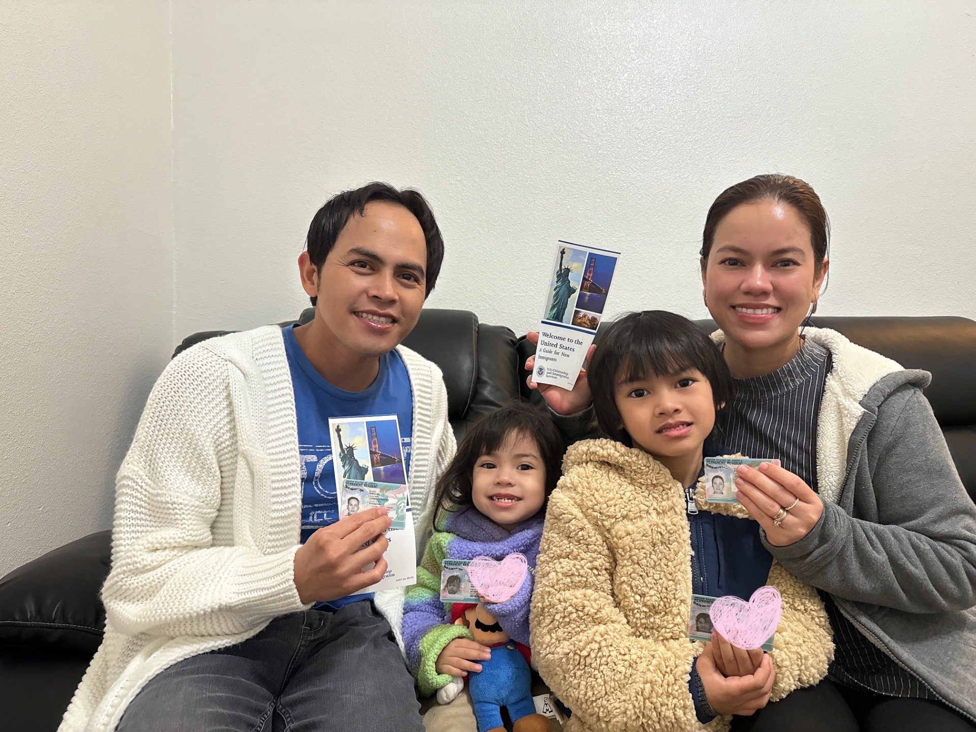 A family of five, including two adults and three children, holds passports and smiles at the camera in a waiting area.