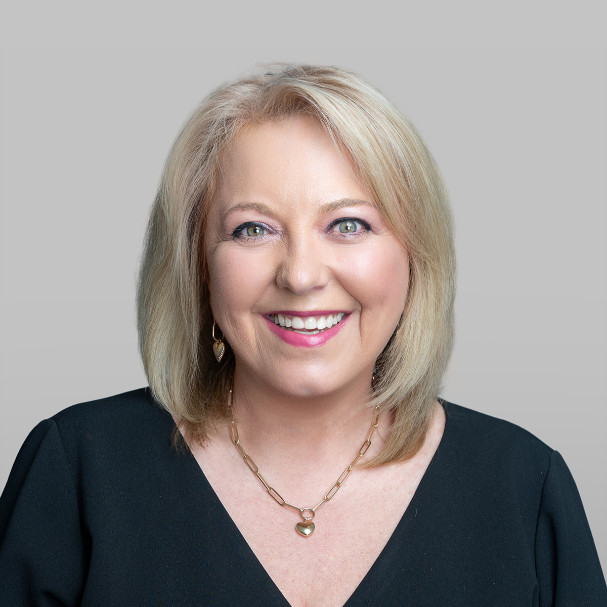 Lesley Hamilton Gray with shoulder-length blonde hair, wearing a black top, gold jewelry, and smiling against a light gray