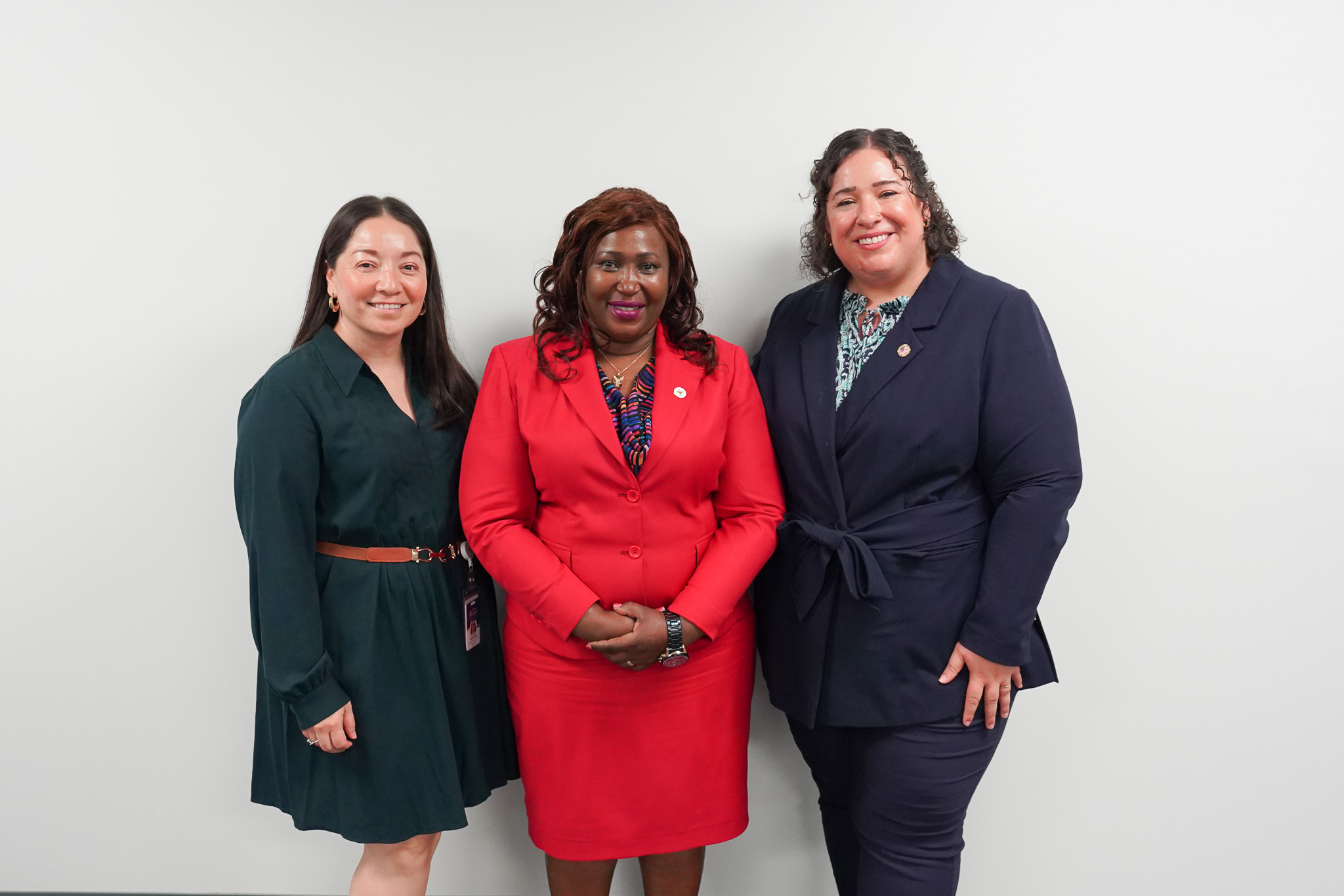 Three women stand together smiling against a plain white wall, dressed in professional attire.