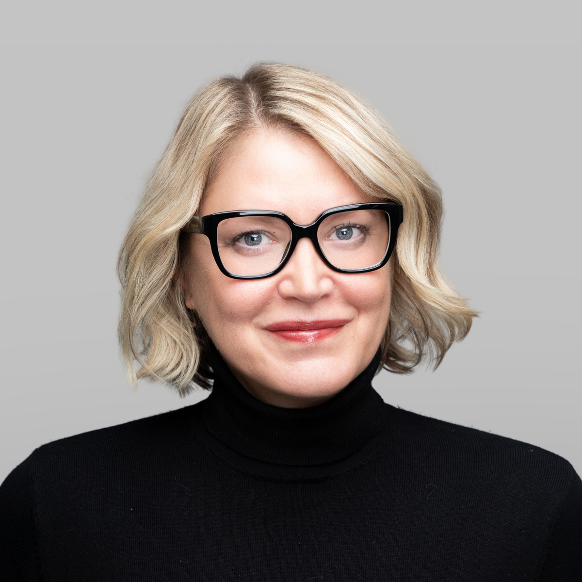 A woman with blonde wavy hair, wearing black-rimmed glasses and a black turtleneck, smiles against a gray background.