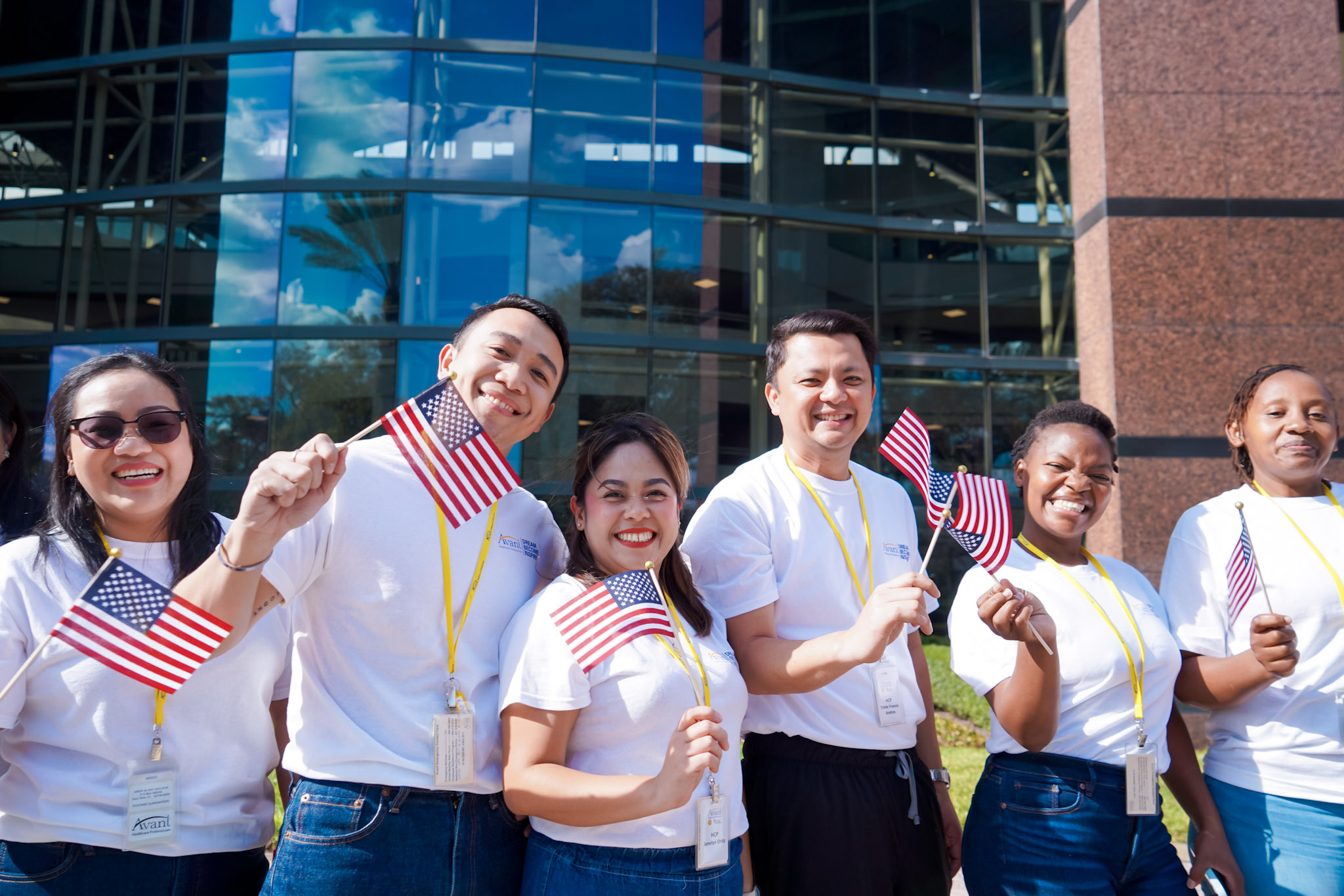 Group of diverse people smiling and waving small American flags outside a modern glass building.