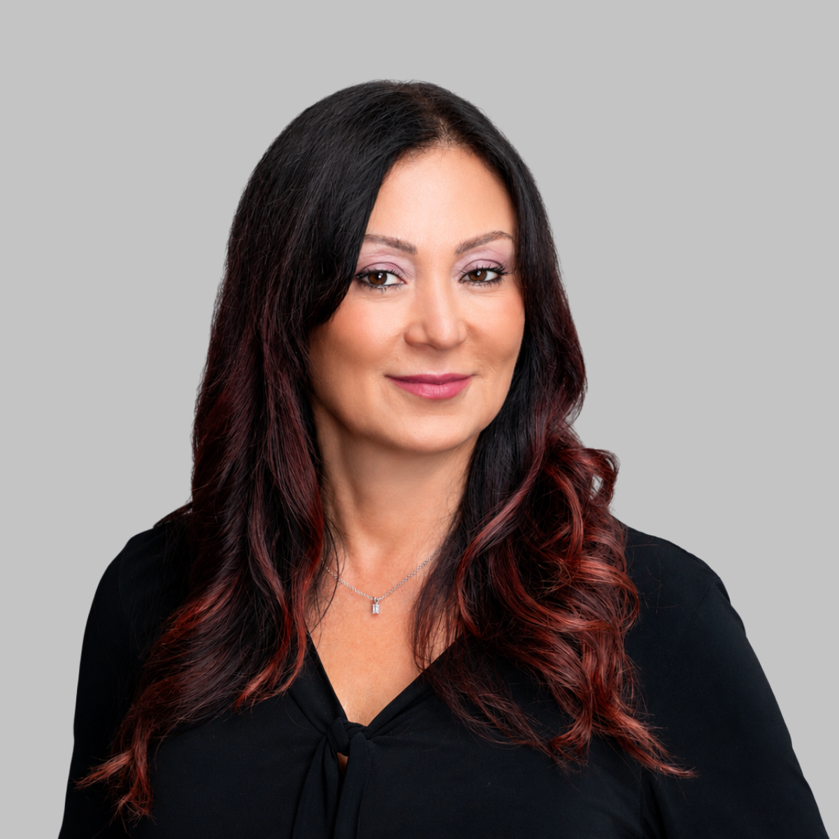 Rebecca Barto with dark hair featuring red highlights, wearing a black top and a delicate necklace, smiling softly against a