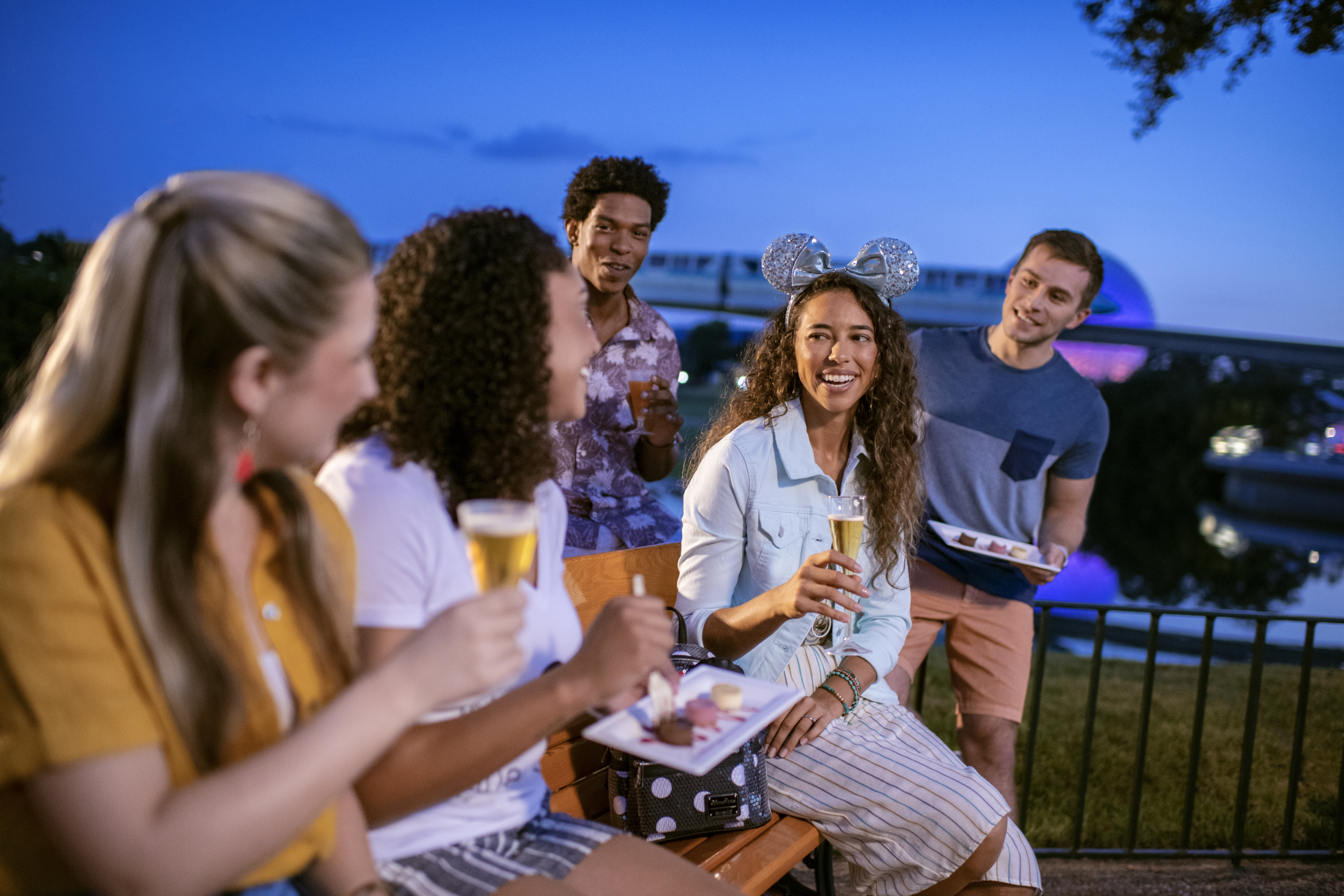 Adults enjoying drinks and snacks at night near EPCOT with Spaceship Earth in the background.