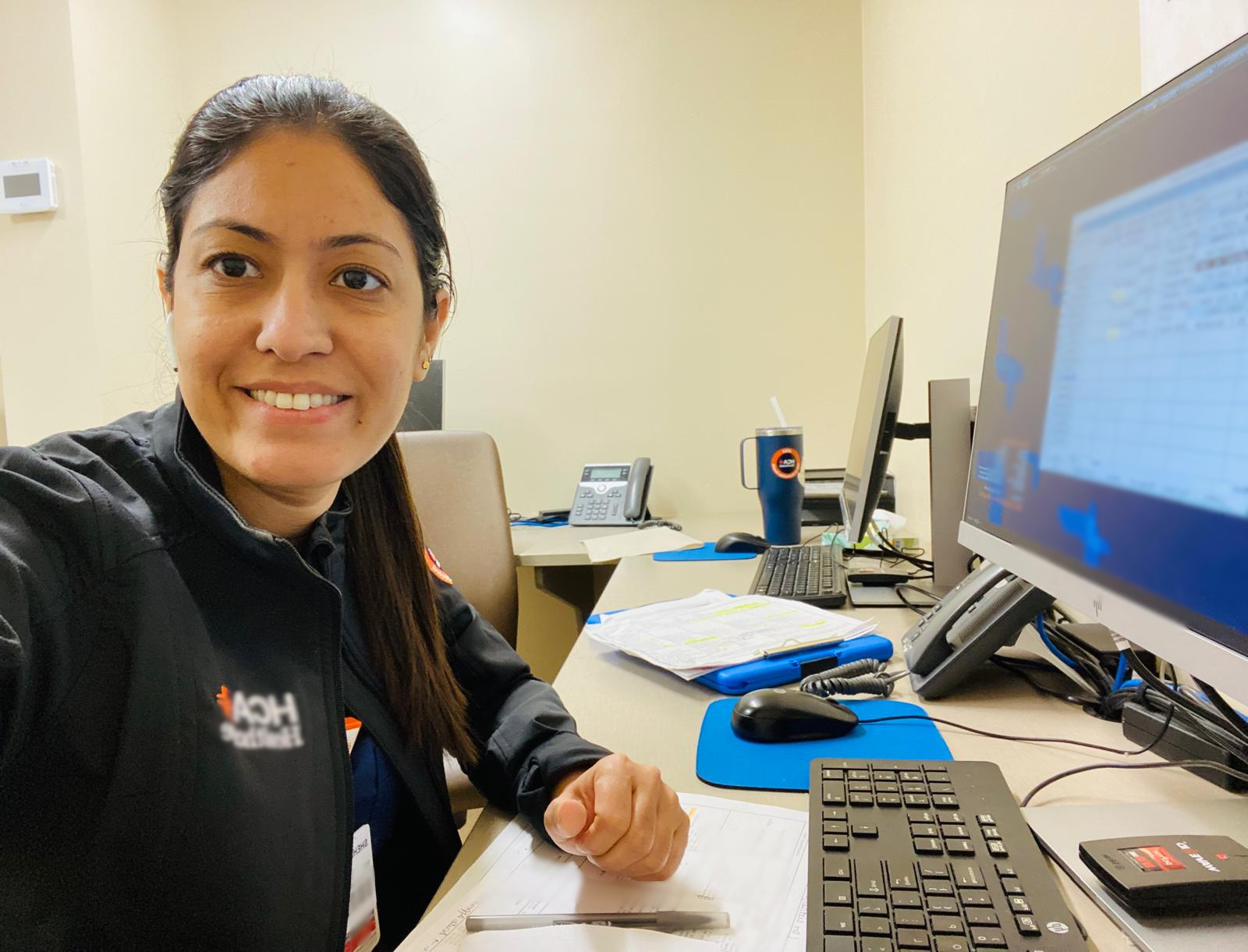 Nurse Shehnaz Ali smiling at her desk with computer, papers, and office supplies in a professional setting.