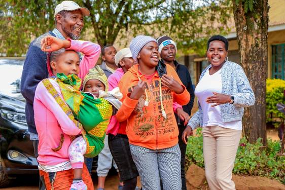A group of smiling people, including children and adults, gather outdoors under trees, enjoying a joyful moment together.