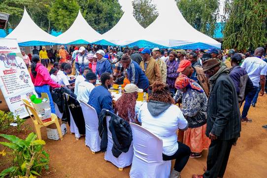 A large group of people gathers outdoors under tents, with some seated at tables and others standing in line.