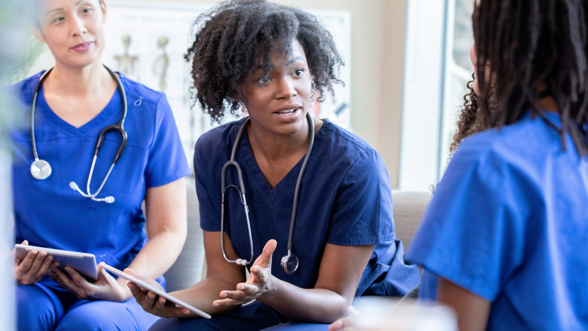 Three diverse healthcare professionals in blue scrubs engage in a discussion, with one explaining something to the others.