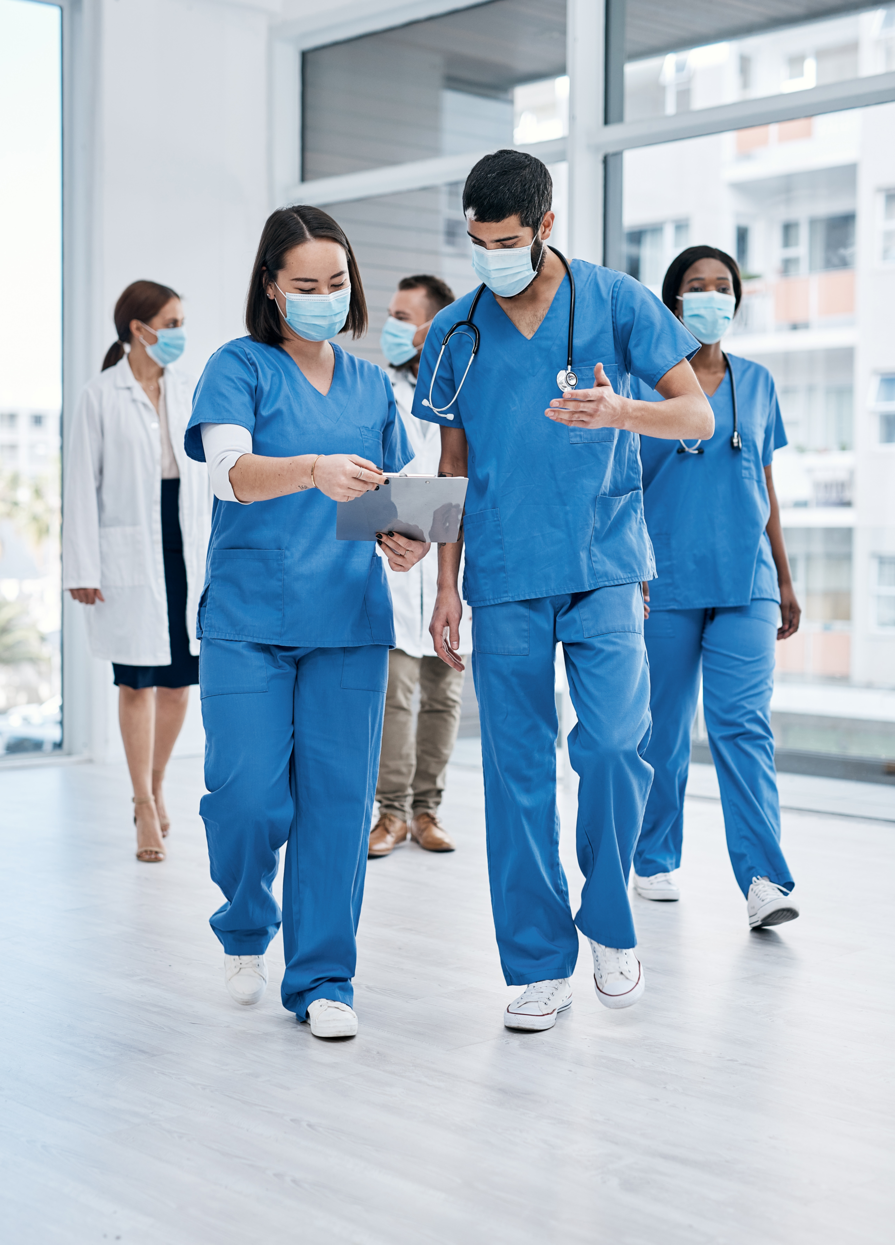 Medical professionals wearing blue scrubs and face masks walk through a hospital corridor.