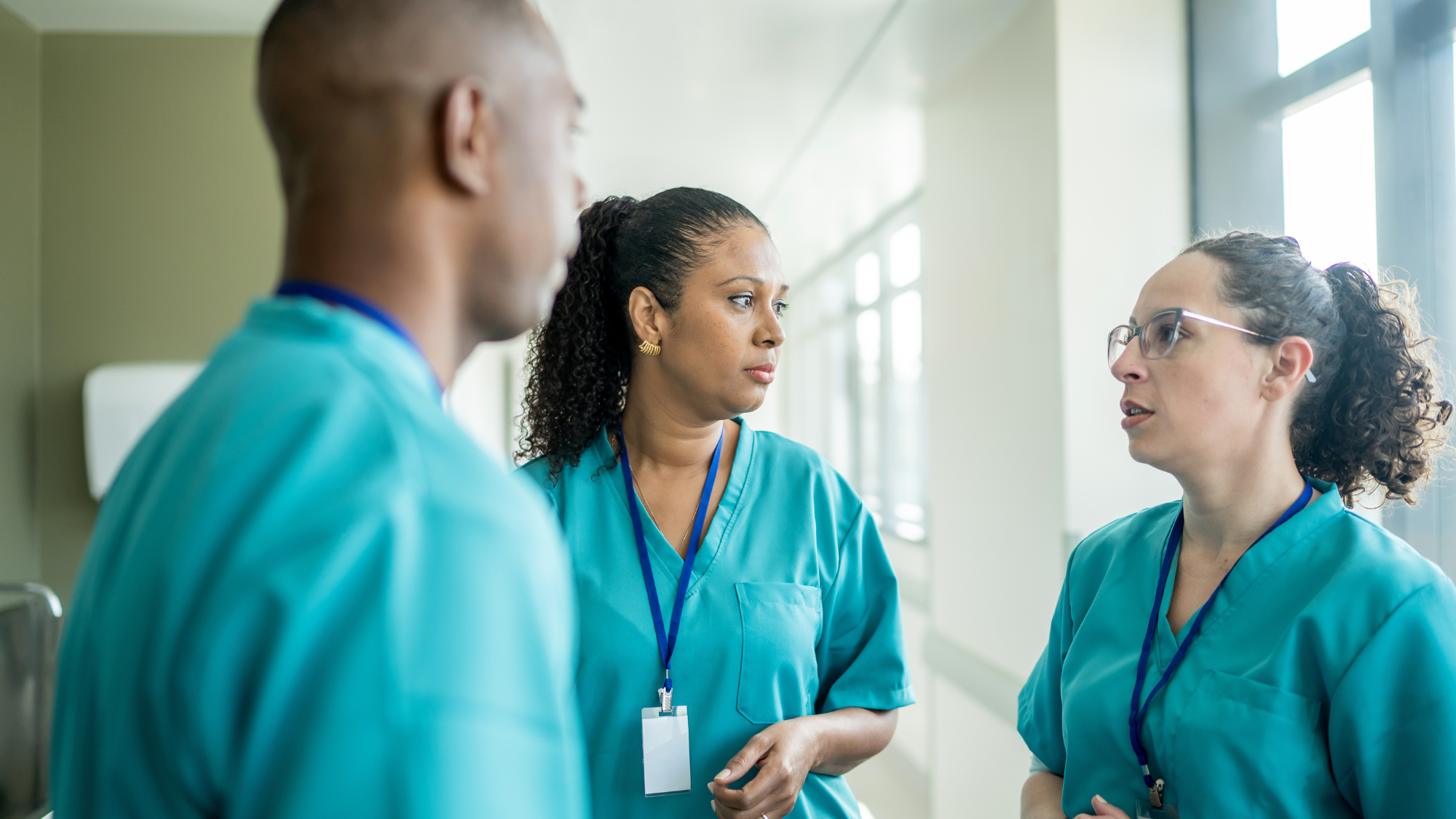 Three healthcare professionals in blue scrubs are engaged in a serious discussion in a hospital corridor.