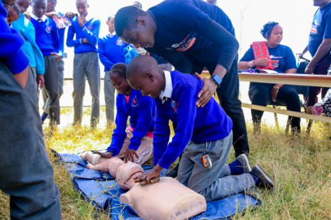 A man teaches children CPR on mannequins outdoors, with students in blue uniforms observing attentively.