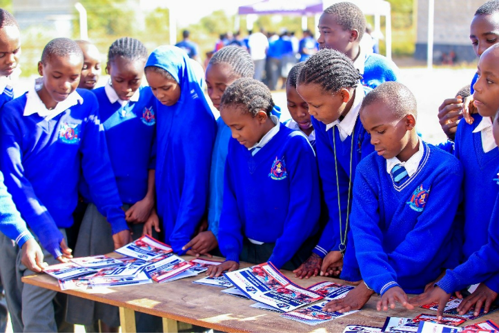 Students in blue uniforms gather around a table looking at printed materials outdoors.