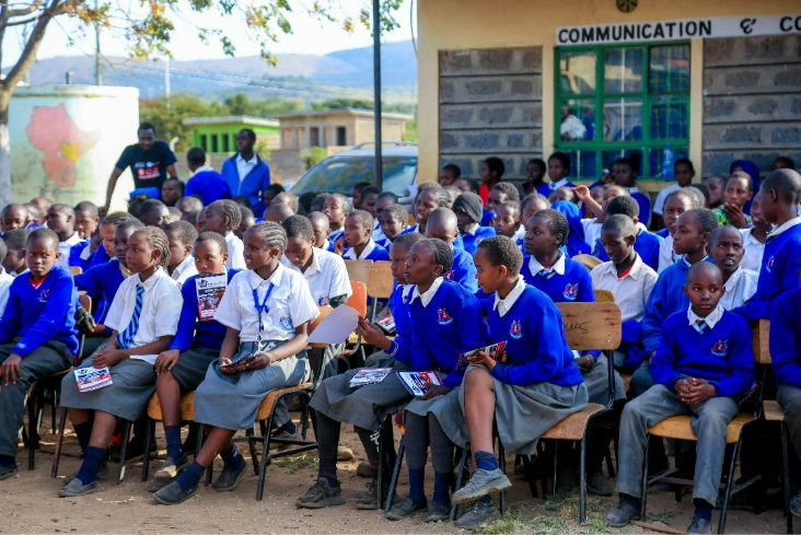 Students in blue uniforms sit on chairs outside a building labeled "Communication," attending a class or event.
