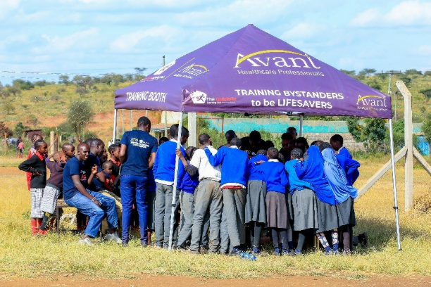 Students gather under a purple tent for a training session outdoors on a grassy field.