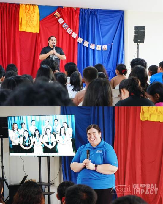 A woman speaks into a microphone in front of an audience, with colorful curtains and a photo display behind her.