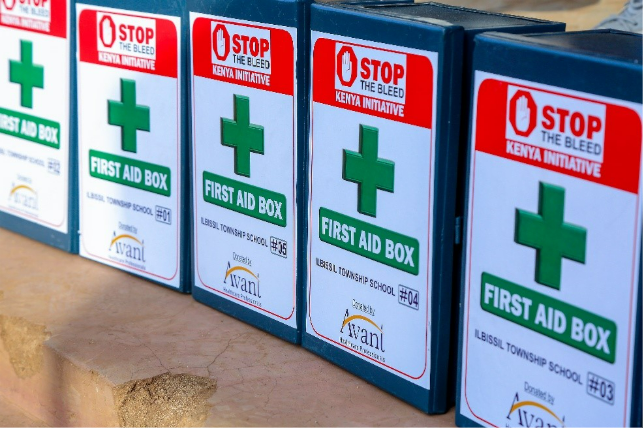 Four first aid boxes labeled "Stop the Bleed" are lined up on a surface, each marked with a green cross and school details.