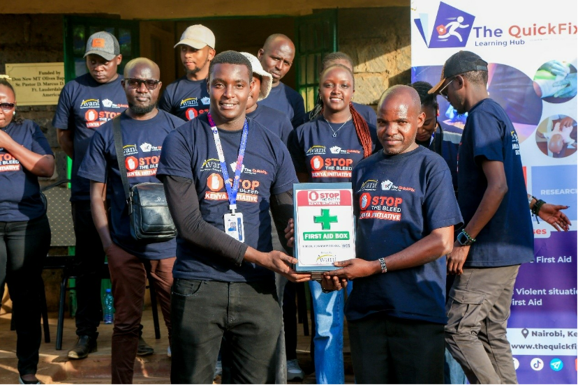 A group of people in navy shirts gather outdoors, with two men holding a first aid box in front of a banner for The QuickFix