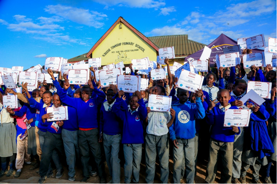 Students in blue uniforms celebrate holding certificates outside a primary school under a bright blue sky.