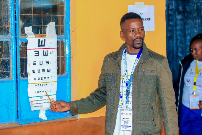 A man points at an eye chart on a blue window, with a woman observing nearby in a room with yellow walls.