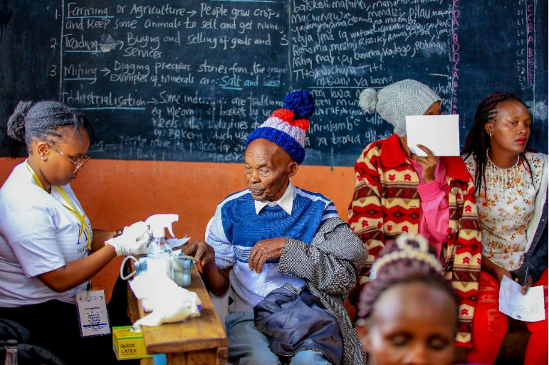 A healthcare worker prepares a man for vaccination while others wait in a classroom with a blackboard in the background.