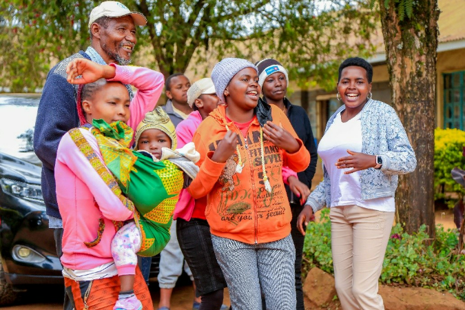 A group of smiling people, including children and adults, gather outdoors near trees and a building, enjoying a joyful