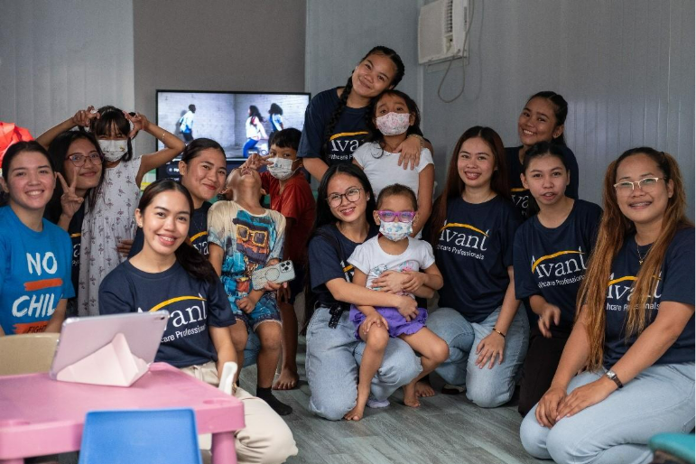 Group of smiling women and children, some wearing masks, gathered indoors with a television in the background.