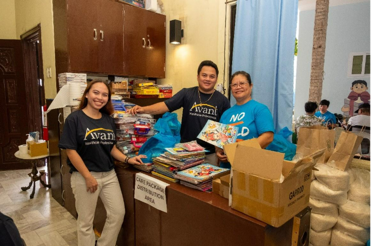 Three people stand behind a table with colorful books and supplies, smiling during a community care package event.