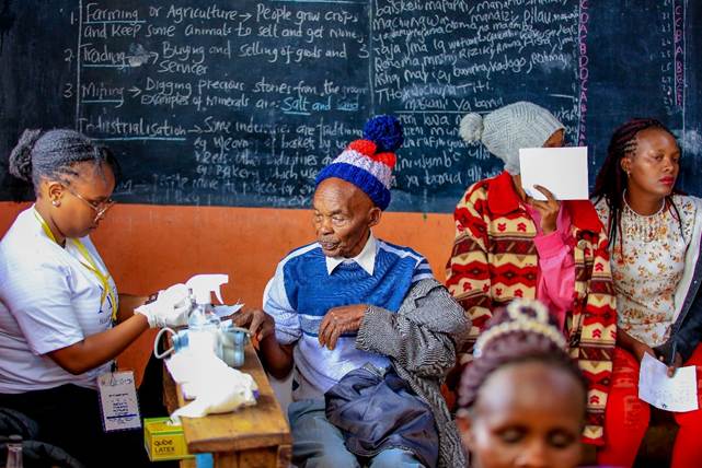 A woman receives a vaccination from a healthcare worker while elderly and other women wait nearby in a classroom with a