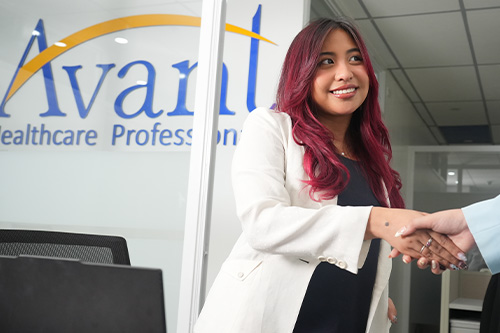 A woman with pink hair in a white blazer shakes hands with someone in an office setting near a sign that reads "Avaril